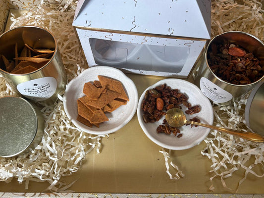 Diwali hamper flat lay: white gift box with gold tins of Whole Wheat Cheese Burst Crackers and Classic Granola Crunch, plated 