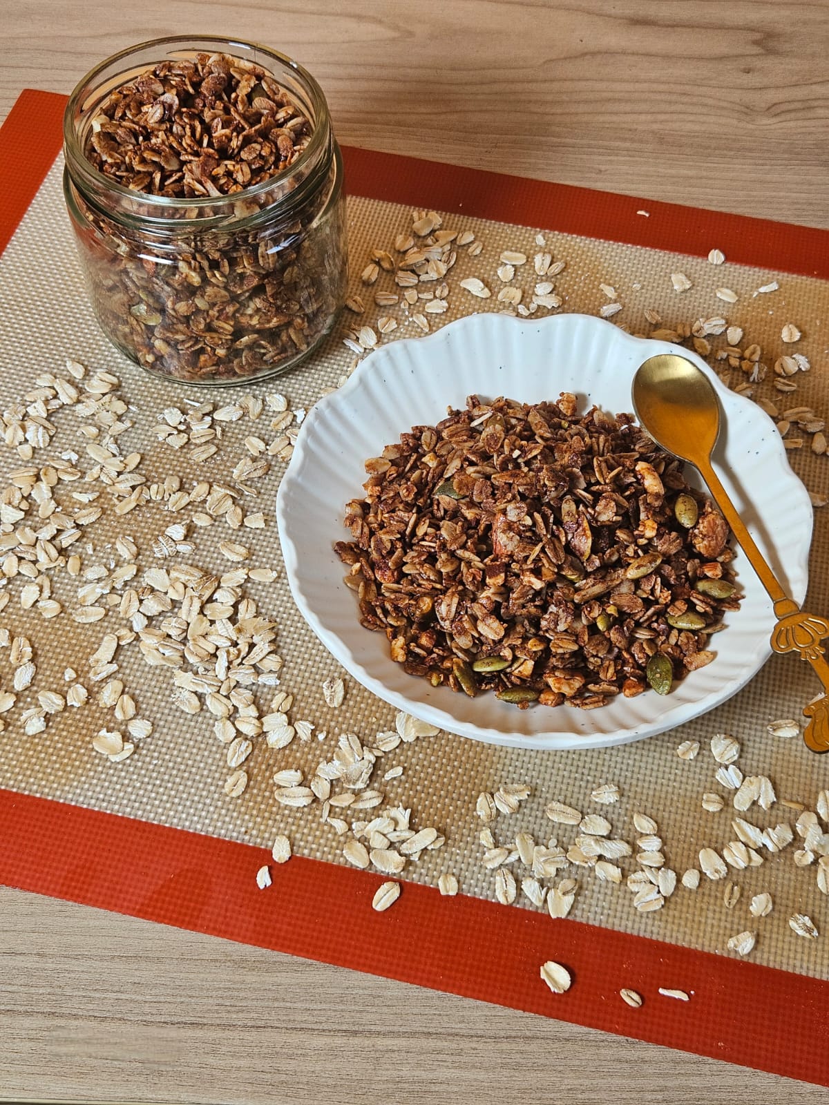 Granola in a glass jar and a white bowl with oats on a baking mat